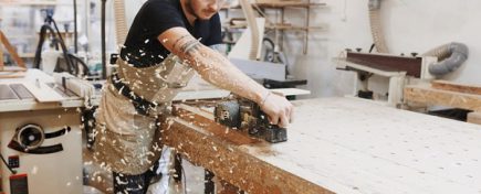 Carpenter working with electric planer on wooden plank in workshop.