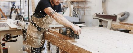 Carpenter working with electric planer on wooden plank in workshop.