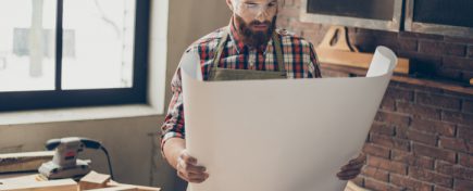 handsome cabinetmaker look at roll of blueprint.  Stylish craftsman with brutal hairstyle and saved glasses work at his workplace