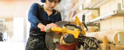 Attractive female carpenter using some power tools for her work in a woodshop